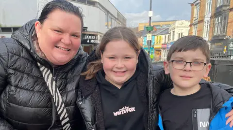 46 -year -old Lisa Mandar is standing on the camera, smiling to the left, a black puff jacket across her shoulder and a zebra print bag. He has pulled his hair back into a ponytail. 10 -year -old daughter Darcy, standing next to her, she is wearing a white fox jumper, which is black and pink text, with black sherpa lining under a black puff jacket, her long hair in a ponytail Has been pulled. The 8-year-old Quad is wearing a black T-shirt and a blue raincoat next to him. He has short hair and is wearing glasses.