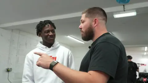 John Nellis Kobbie Mainoo wearing a white jumper and John Nellis wearing a black top, John is pointing