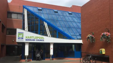 BBC Front of Hartlepool Borough Council HQ, showing the main entrance, a blue metal and glass atrium, with brick walls to the side, the right hand one featuring two hanging baskets.