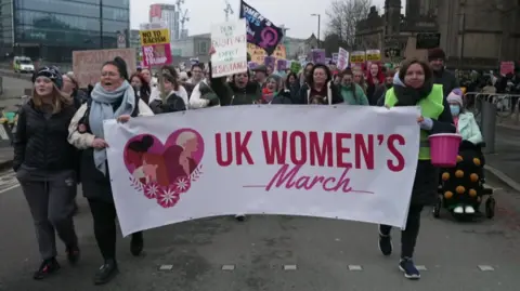 Women hold a large white and pink banner saying UK Women's March while marching near Manchester Cathedral