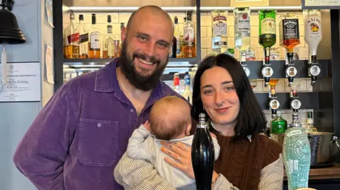 Three people including a baby standing at the bar of a pub. Behind them are rows of bottles of alcohol and some on optics.