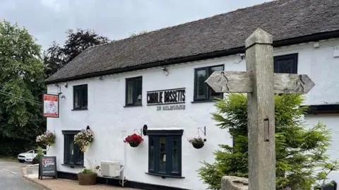 Laura Ball The black and white pub is pictured from the road. It has black-framed windows and a small tree growing on one side.