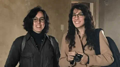 Peregrine Bryant Two young women smile at the camera. One with shorter brown hair and glasses wears a dark coat and straps showing she has a rucksack on. Next to her is a longer haired brunette woman wearing a light brown coat holding a camera. They are standing in a room with beige plaster walls.