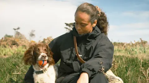 Sam Fernandes A woman wearing a black coat and black trousers. She has brown curly hair that is tied back with a pink scrunchie. There is a small brown and white dog next to her with a orange ball in her mouth. They are sat on grass. The sky is blue with white clouds. 