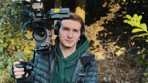 Luke Jones A young man with swept back brown hair holding a large camera kit on his shoulder. He is standing outside next to a bush and is wearing a dark green hoodie and waterproof coat.