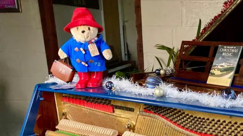A Paddington Bear teddy, in red wellies, blue duffle coat and red hat, standing on top of a blue piano. A book of "Christmas Music" is on a music stand on top of the upright piano. The piano has no front on it, and the hammers, etc. are visible.