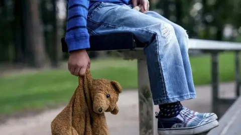 Getty Images The picture appears to be a primary school aged boy sitting on a bench and dangling a saggy brown teddy bear from his right hand. The boy, who is only visible from the waist down, is wearing a blue long sleeved top with dark blue stripes and light blue jeans with rips across the knees. He also has dark blue socks with a white pattern and blue and red tartan slip on pumps