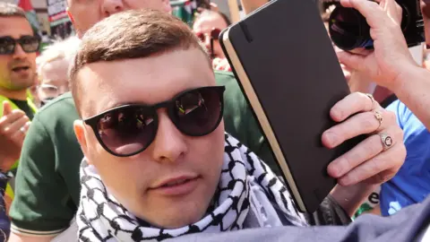 PA Media A close-up of a man in the midst of a crowd. He is wearing sunglasses and a black and white keffiyeh headdress around his neck like a scarf. There is a camera lens being held close to his face as a photographer takes a photo