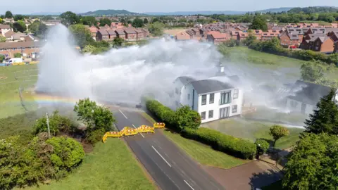 A drone shot of the burst water pipe in Matson, it shows a large torrent of water shooting from a field, across a two lane road and directly onto a large house. The road has been cordoned off with yellow fencing.