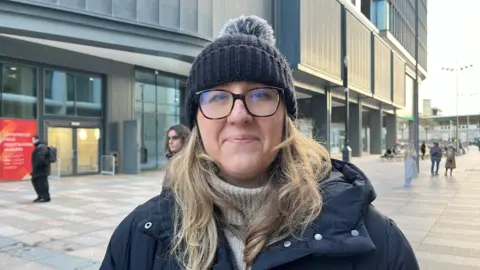 Gabriella Lake, in a coat, hat and glasses looking at the camera