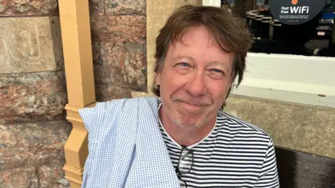 A man wearing a striped top, with a blue oxford shirt hanging over his right shoulder and glasses hanging from his collar. He is smiling at the camera infront of a brick wall, sitting on a bench.
