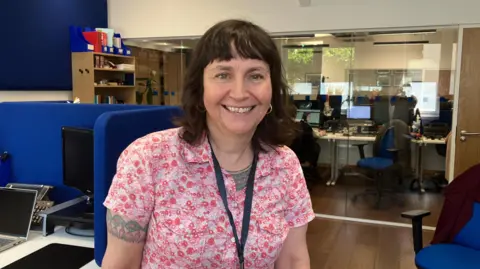 Kit Taylor/BBC A woman with brown hair and a pink flowery top in a helpline office, with sectioned off booths and computers. 