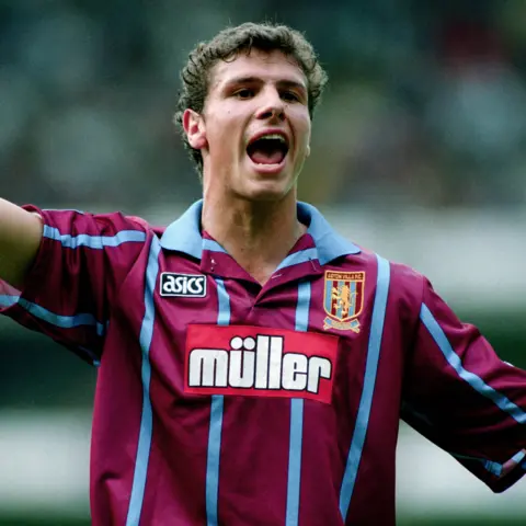 Offside via Getty Images A young man shouting and pointing during a football match. He has short, bushy brown hair and is wearing a claret football shirt with light blue stripes and trim.