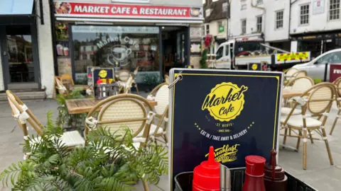 A sign for Market Cafe with chairs and tables and plants on a paved area behind it and the cafe itself beyond that. 