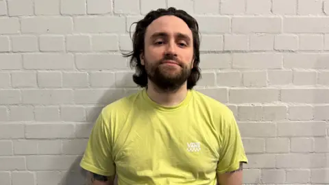 Josh Henderson A man with mid length dark hair and a yellow top looking at the camera.