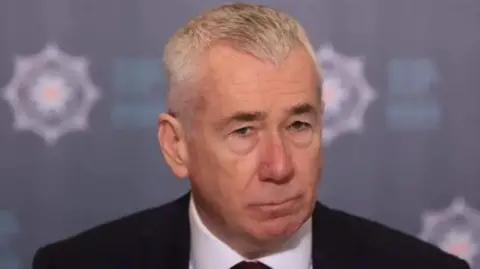 PA Media Jon Boutcher. He has white hair, wearing a black blazer, white shirt and dark tie. PSNI signage is in the background.