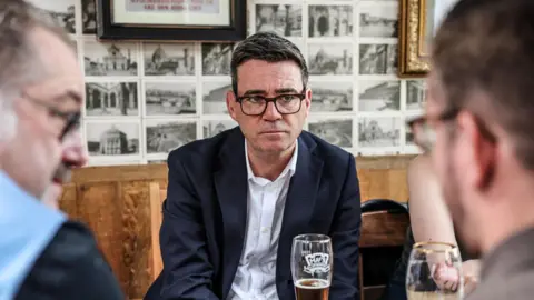Reuters Andy Burnham has dark hair and is wearing dark glasses. He is wearing a white shirt with a dark suit jacket and is sitting at a table with a pint in front of him. He is facing two men who are talking