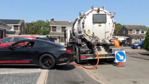 A sewage tanker parked across a driveway as a black car is trying to reverse off.