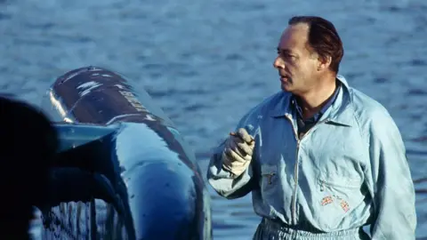 Getty Images Donald Campbell, standing next to Bluebird. He has dark hair, brushed back from his face, and is wearing light blue overalls.