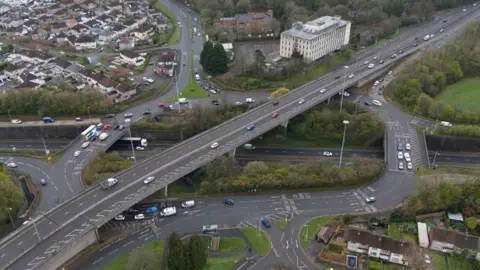 An aerial view of Manadon Roundabout which shows one large road passing over the roundabout with cars travelling around it. 