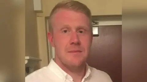 A man in a light coloured shirt and short hair with a door behind him.