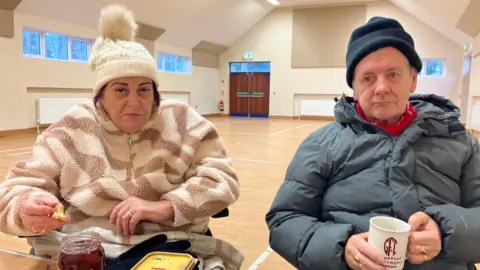 Desmond and Olive Hyndman sit in a community centre. Desmond holds a mug and Olive holds a scone. There is butter and jam in front of them. Desmond wears a grey puffer jacket and grey hat and Olive wears a fleece and cream hat.