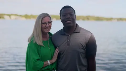 Liam Weir/BBC Stacey and Marven facing the camera in front of a lake. Both are smiling, Marven is wearing a grey, collared T-shirt, Stacey has long blond hair and glasses and is wearing a bright green gathered top.