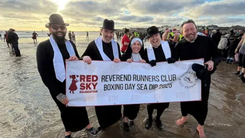 Jim Scott/BBC Un grupo de cinco personas vestidas de vicarios, con túnicas negras y accesorios blancos. La mujer del centro lleva un atuendo de monja. Sostienen una pancarta que dice: «Reverendo Club de Corredores, Boxing Day Sea Dip 2025».