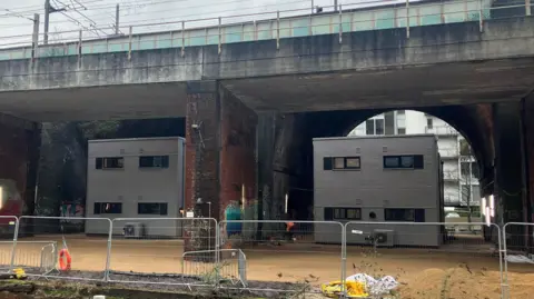 Photograph of the modular homes which are being built underneath a serires of railway arches in Castlefield. The Bridgewater Canal can be seen in the foreground, surrounded by metal fencing.