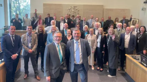 Reform UK A large group of people in suits with the men wearing light blue ties in a council chamber.