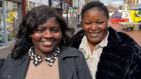 Two women are standing on the high street facing the camera and smiling. The woman on the left has dark hair, gold earrings a leopard print collar and grey coat. The woman on the right ha dark hair, a god necklace and is wearing a black furry coat.