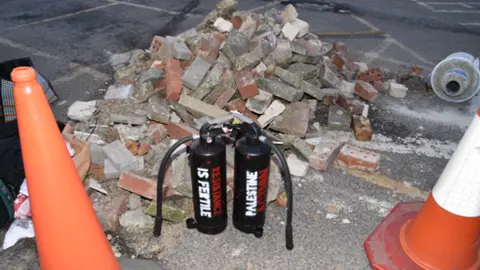 CPS Two black fire extinguishers stand in front of a pile of bricks and rubble. One has "resistance is fertile" in red and white letters and the other "Palestine action" written on.