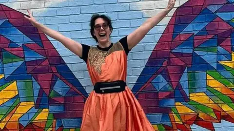 Jennifer Gane is wearing an orange and gold ballgown. She has her hands raised in the air and a bright smile on her face. She is standing in front of a colourful mural which makes it look like she has wings.
