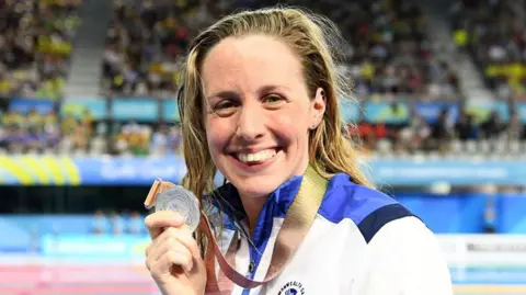 Getty Images Swimmer Hannah Miley grins while holding a silver medal won at Glasgow 2014. She has wet blonde hair and is wearing a blue and white Team Scotland tracksuit