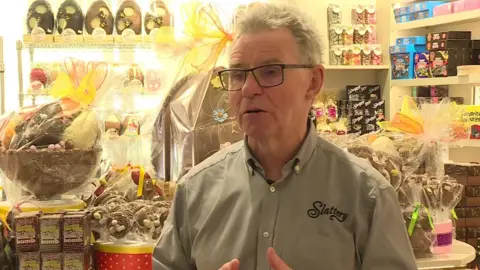 BBC John Slattery, standing in front of lots of chocolate. He has grey hair, is wearing glasses and a light buttoned shirt with Slaterry printed on.