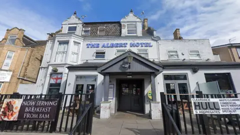 Google A Google Street View of the exterior of The Albert Hotel. It shows a white building with blue letters above the entrance reading "The Albert Hotel". There are stairs and black railings leading up to the entrance of the hotel. The sky is blue and cloudy. 