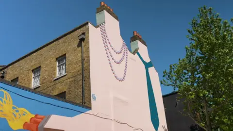A mural of a string of pearls and a tie on the side of a building. The mural uses two chimneys on the building to create the illusion of necks.