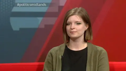 A young woman in a TV studio. She has short cropped hair and is wearing a green cardigan.
