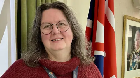 Local Democracy Reporting Service Councillor Charmaine Morgan - a woman with shoulder-length grey hair with round thin black-rimmed glasses - is smiling at the camera. She is wearing a red turtle neck jumper and a lanyard. She is standing next to a green curtain, a union jack flag and a photo of the king.
