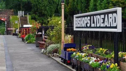 Supplied The platform at Bishop's Lydeard with a sign on the right of the shot with the name of the station. There are flowers running along the edge of the platform as well as a shelter further down. In the background are trees surrounding the entire platform.