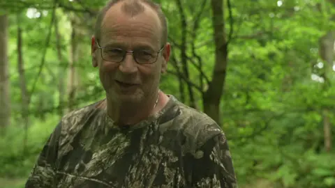 A man with very short hair, wearing glasses, a silver necklace and a green patterned T-shirt, is looking to the right. Behind him is a forested area.