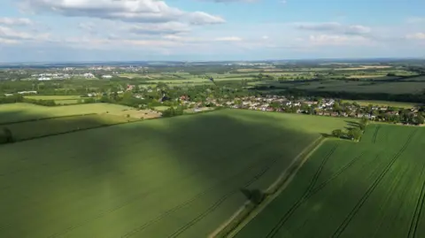 Anna Gazeley An aerial view of green fields with small residential settlements visible in the distance. 