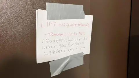George Carden/BBC A handwritten sign stuck to the brown lift door with duct tape saying the engineer didnt have parts to repair the lift