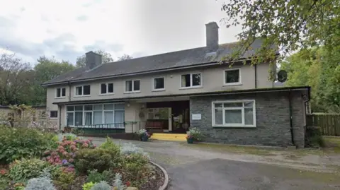 A long 1970s style building made of concrete sits at the top of a short curving drive. It is two storeys and has several windows with shrubs to the left hand side of the drive and trees behind. 