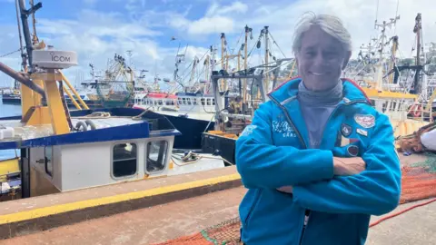 Ian Perkes is smiling and has his arms crossed as he stands on the quay at Brixham harbour. He has quite long grey hair and is wearing a sail racing logo's jacket. The boats behind him are fishing vessels.