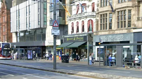 Google Princes Street in Edinburgh - crowds of people walk along the pavements while buses pick up passengers from bus stops.