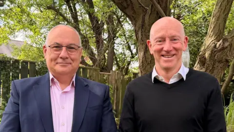 Supplied Edward Gorringe and Mike Thornicroft stand side-by-side in a garden and are both smiling at the camera. Gorringe has balding white hair and glasses. He is wearing a blue suit and a pink shirt. Thornicroft is bald and is wearing a black jumper and a white shirt.