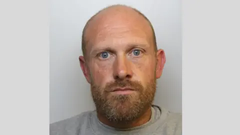 Avon and Somerset Police Police mugshot of James Stock from custody. He is wearing a light gray jumper and has a bald head, blue eyes and a ginger beard.