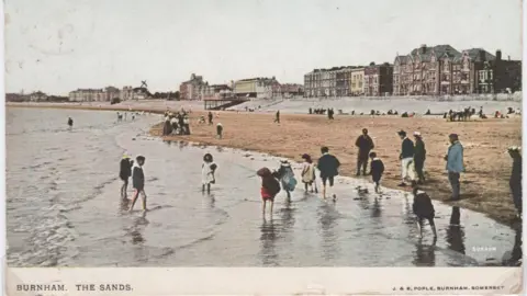 North Sedgemoor Local History Group An old postcard showing Burnham's beach. People are walking through the water. In the background are houses along the promenade.