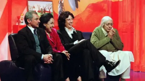 A photo taken from the show The Kumars at No 42, showing four people sat on a sofa in front of an orange curtain. The picture Shows Vincent Ebrahim, Indiri Joshi, Laurence Llewelyn Bowen and Meera Syal. Syal is dressed up as an old woman.